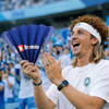 A person with curly hair holds a blue Clap Banner, smiling enthusiastically amidst a cheering crowd.