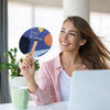 A round wooden paper fan with a colourful design, held by a smiling woman in a casual setting. A logo is present.