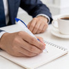 A hand holds a blue and white pen over an open notepad, with a cup of coffee in the background.