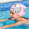 A silicone swim cap in pink with a fish design, featuring a large eye and sharp teeth, worn by a girl in a pool.