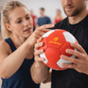 A hand-stitched handball in red and white, held by a man and a woman, with a logo displayed on its surface.