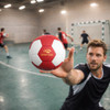 A red and white thermal bonded handball is being held by a player in an indoor sports setting, with teammates in the background.