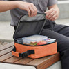 An orange cooler bag with a zippered opening, containing food containers and an apple, placed on a wooden bench.