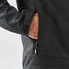 A black softshell jacket with a zippered pocket, featuring a hand reaching inside the pocket.