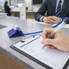 A blue fixed signature pen with a clear grip is placed on a grey countertop beside a clipboard and a hand writing.