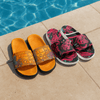 Two pairs of slides sit by a pool: one in orange with a patterned strap, the other in black with a floral design.