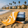 A stack of five bath towels in various colours, including yellow, blue, and black, on a sandy beach with palm trees and houses in the background.