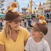 A brown baseball cap featuring a plush bird on top, worn by a woman and a child at a festive outdoor location.