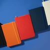 A collection of Minichroma A6 Notebooks in black, orange, red, navy, and white, arranged on a blue surface.