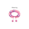 A pink crochet pet bib features scalloped edges, purple and white flower embellishments, and tied ends with pom-poms.