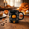 A black ceramic coffee mug featuring Halloween-themed designs, with a pumpkin centrepiece and a logo.
