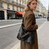 A black polyurethane tote bag is being carried by a model dressed in a brown coat on a Parisian street.
