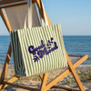 A green and white striped tote bag with a branding logo sits on a wooden beach chair, with a scenic ocean view in the background.