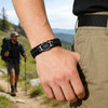 A braided paracord bracelet in black with a themed pendant worn on a hand, outdoors in a mountainous setting.