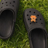 A pair of black clog shoes featuring a 3D bear charm on one shoe, set on green grass.