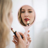 A pink handheld makeup mirror reflects a person applying lipstick, with a soft, neutral background.