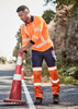 A man in high-visibility orange and navy work pants adjusts a traffic cone on a road. The pants feature reflective strips.