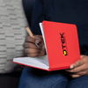 A red A5 notebook with a logo on the cover, held open with a hand writing inside.
