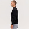 A black crewneck sweatshirt is worn by a model standing sideways, paired with grey sweatpants. The sweatshirt has a logo.