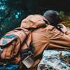 A person in a brown jacket with a backpack gazes into the distance, surrounded by nature.