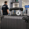A round white luggage tag attached to a black suitcase at an airport security checkpoint, featuring a logo.