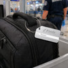 A square brushed aluminium luggage tag attached to a black bag, featuring a logo and a name label.