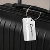 A square aluminium luggage tag in silver attached to a black suitcase, featuring a logo and a name label.