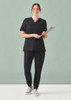 A woman wearing black women's Pulse jogger scrub pants and a matching top, holding a clipboard, standing against a light green background.