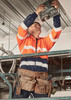 A worker in an orange and navy hi-vis full zip hoodie uses a tool while standing on a scaffold, wearing a hard hat.
