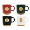 A set of four ceramic coffee mugs in red, green, black, and white, each with a gold medal on the base and a logo.