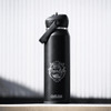 A drink bottle in matte black with a flip straw lid and a logo featuring a vanlife theme.