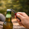 A stainless steel multi-function utensil opening a green drink bottle on a wooden surface in an outdoor setting.
