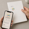 A person holding a phone displaying a coffee shop profile next to a white notebook with a colourful sticker.