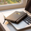 A5 weekly diary in black with gold initials, resting on a laptop keyboard by a sunlit window.