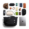 A black messenger bag with various items including sunglasses, a hat, a laptop, and a wallet, arranged on a flat surface.