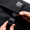A black messenger bag with a flap and fastening mechanism, featuring a logo. A hand is holding the clasp.