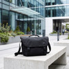 A black satchel bag sits on a concrete bench in a modern urban setting with greenery. It features a logo.