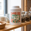A white tea canister with green illustrations of two cheerful mugs, displayed on a wooden shelf alongside jars and cups.