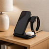 A black 3 in 1 wireless charger for a phone, earbuds, and a smartwatch, placed on a wooden table beside a lamp.