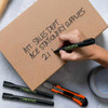 A hand writes on a cardboard box using a black Staedtler Lumocolor permanent marker with a chisel tip.