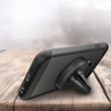 A black round car phone holder positioned behind a smartphone on a wooden surface. The holder has a logo.
