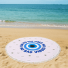 A round beach towel in white and blue with a central eye design, placed on a sandy beach by the water.