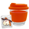 A clear glass coffee cup with an orange silicone lid and sleeve, accompanied by a bag of mixed jelly lollies.