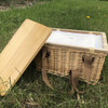 A square wicker picnic cooler basket in natural tones with a bamboo lid and brown straps, placed on grass.