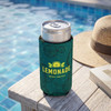 A slim can cooler in dark green with a logo, holding a chilled lemonade drink, placed on a wooden table by a pool.