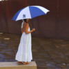 A blue and white umbrella with a logo, held by a person in a white dress, standing near water.