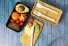 A two-layer lunch box in black and beige contains utensils and food items like fruit, bread, cheese, and vegetables.