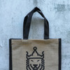 A jute tote bag in natural brown with black handles and a lion design in the centre. It features branding on the side.
