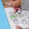 A hand colouring in a bear illustration on a notepad with a pink pencil, showcasing vibrant pastel colours.