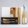 A wooden cheese board, a two-piece gold knife set in a black box, and a bottle of white wine with a colourful label.